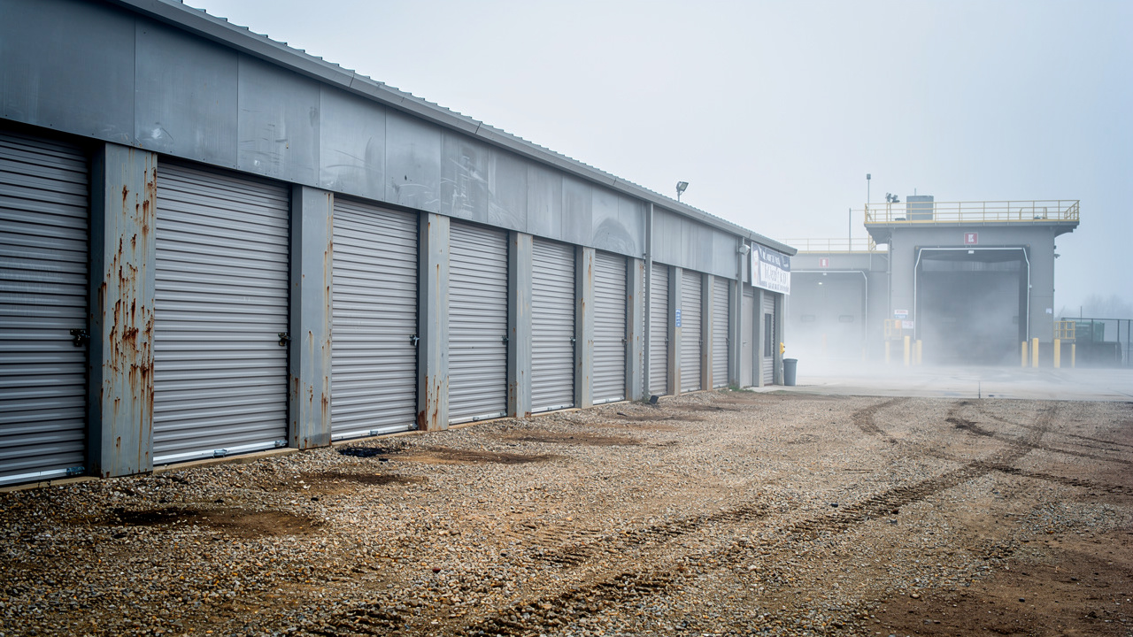 Des conteneurs de stockage en acier alignés sur un site industriel, à côté d’un entrepôt, avec une cour gravillonnée et une lumière de matin.